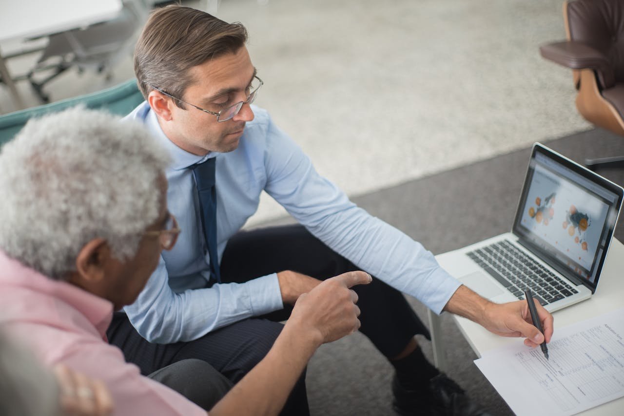 Two men in a business meeting reviewing financial documents and discussing data on a laptop.